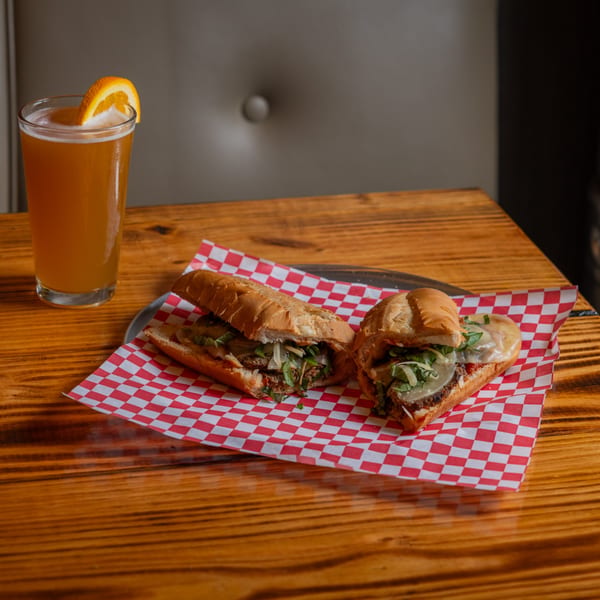 Two hearty sandwiches on red and white checkered paper with a beer garnished with an orange slice.