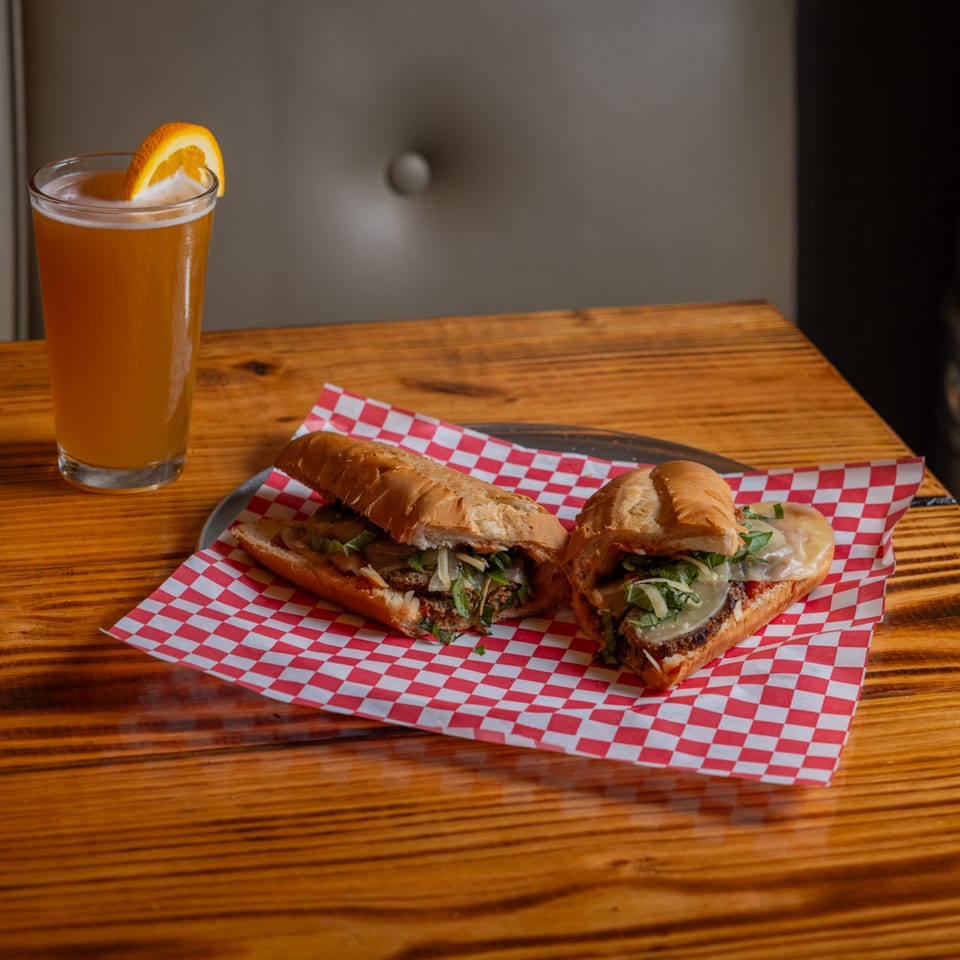 Two hearty sandwiches on red and white checkered paper with a beer garnished with an orange slice.
