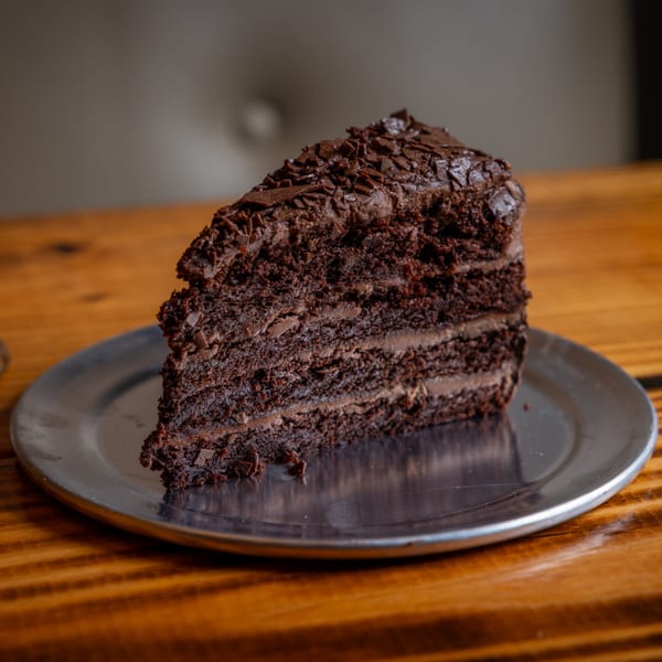 A rich slice of chocolate cake with chocolate frosting and shavings on a silver plate.