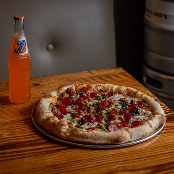 A pepperoni, sausage, olive, and onion pizza on a wooden table with a Fanta soda.
