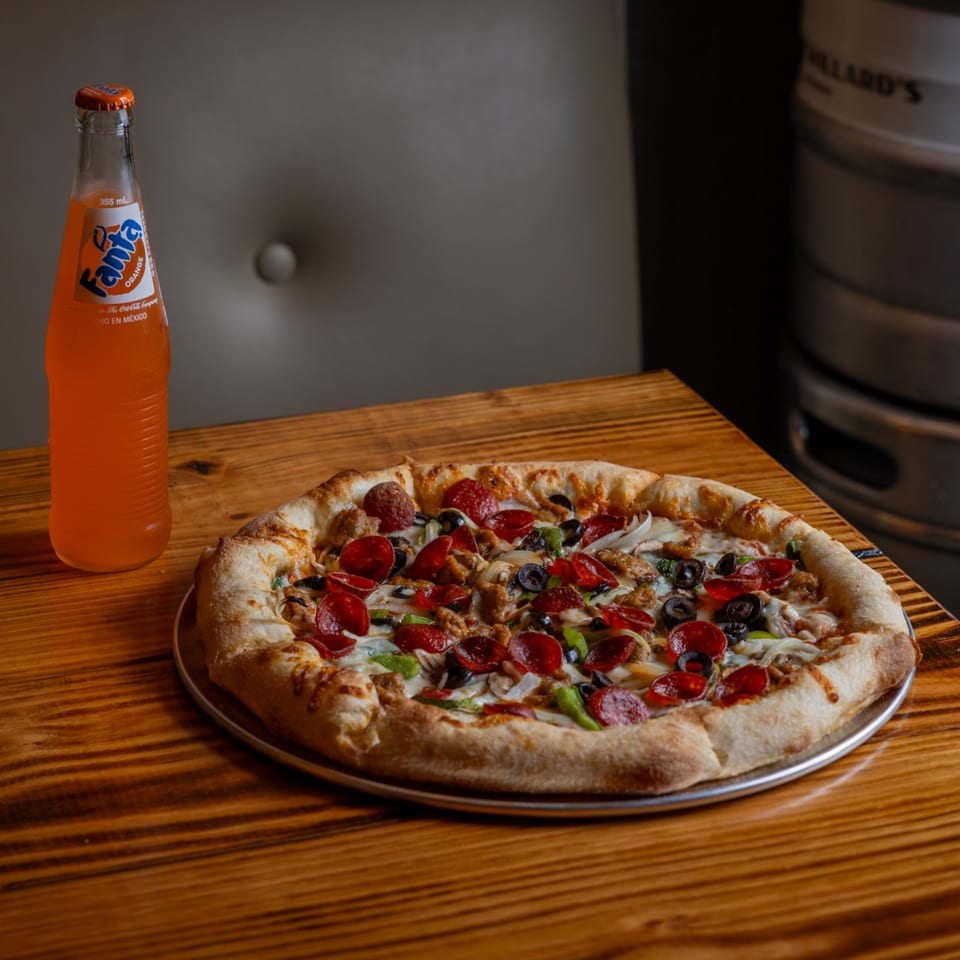 A pepperoni, sausage, olive, and onion pizza on a wooden table with a Fanta soda.