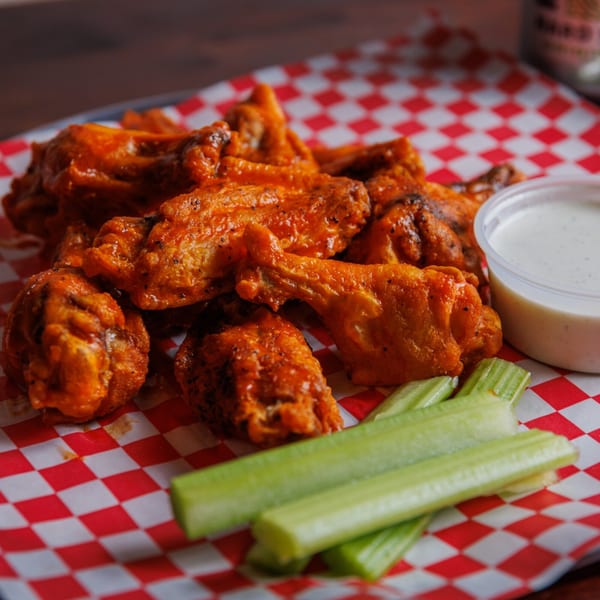 A platter of buffalo wings with celery sticks and ranch dressing on red and white checkered paper.